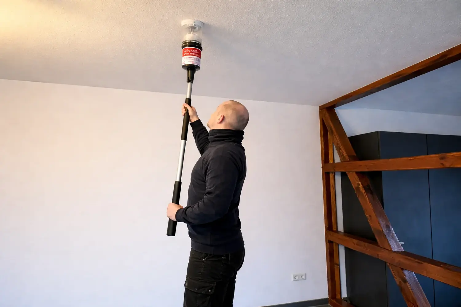 Brandschutz Tobias Scheunemann - Brandschutz und Schulung - Ein Mann benutzt eine Verlängerungsstange, um einen an einer weißen Decke installierten Rauchmelder zu testen.