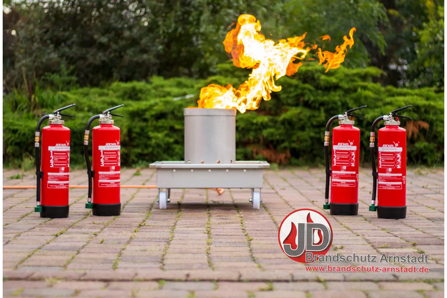 Brandschutz Tobias Scheunemann - Brandschutz und Schulung - Eine Übungsanlage für Brandschutzmaßnahmen im Freien mit einem kontrollierten Feuer in einem Metallbehälter, flankiert von zwei roten Feuerlöschern auf Pflastersteinen.