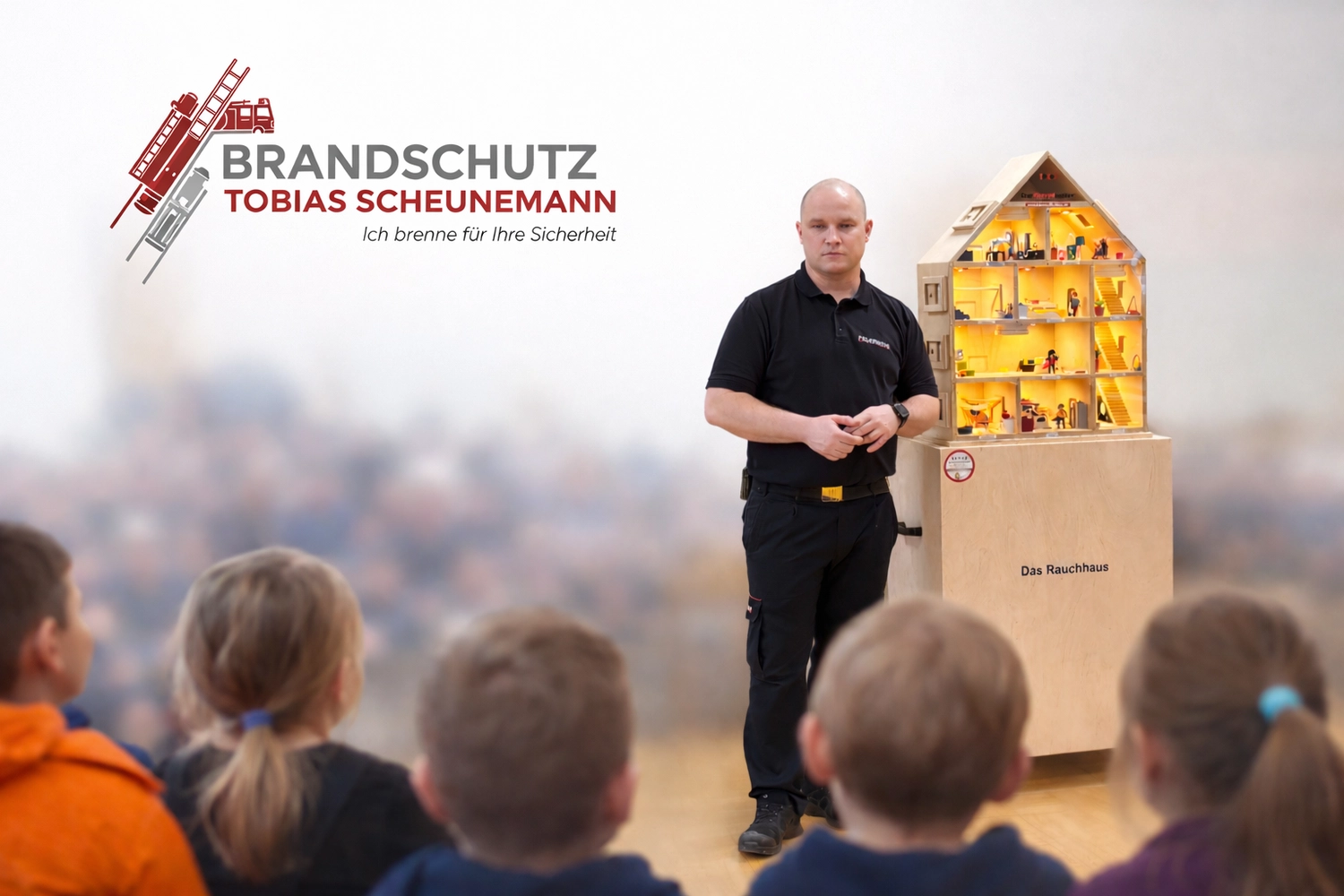 Brandschutz Tobias Scheunemann - Brandschutz und Schulung - Ein Feuerwehrmann steht neben einem Modellhaus auf einem Podest und gibt einer Gruppe junger Kinder, die auf dem Boden sitzen, eine Präsentation.